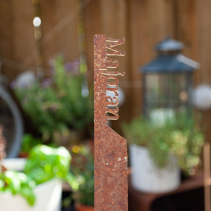 Kräuterstecker ‚Majoran‘ aus Metall in Rostoptik, freigestellt im Garten mit unscharfem Hintergrund.
